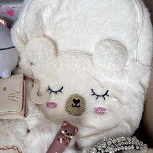 Puede incluir: Una mochila blanca y esponjosa con un dise&ntilde;o de cara de oso, con ojos cerrados, detalles de purpurina rosa y una nariz de purpurina dorada. Otros art&iacute;culos incluyen una cartera peque&ntilde;a, un labial marr&oacute;n con dise&ntilde;o de oso y una diadema de perlas.