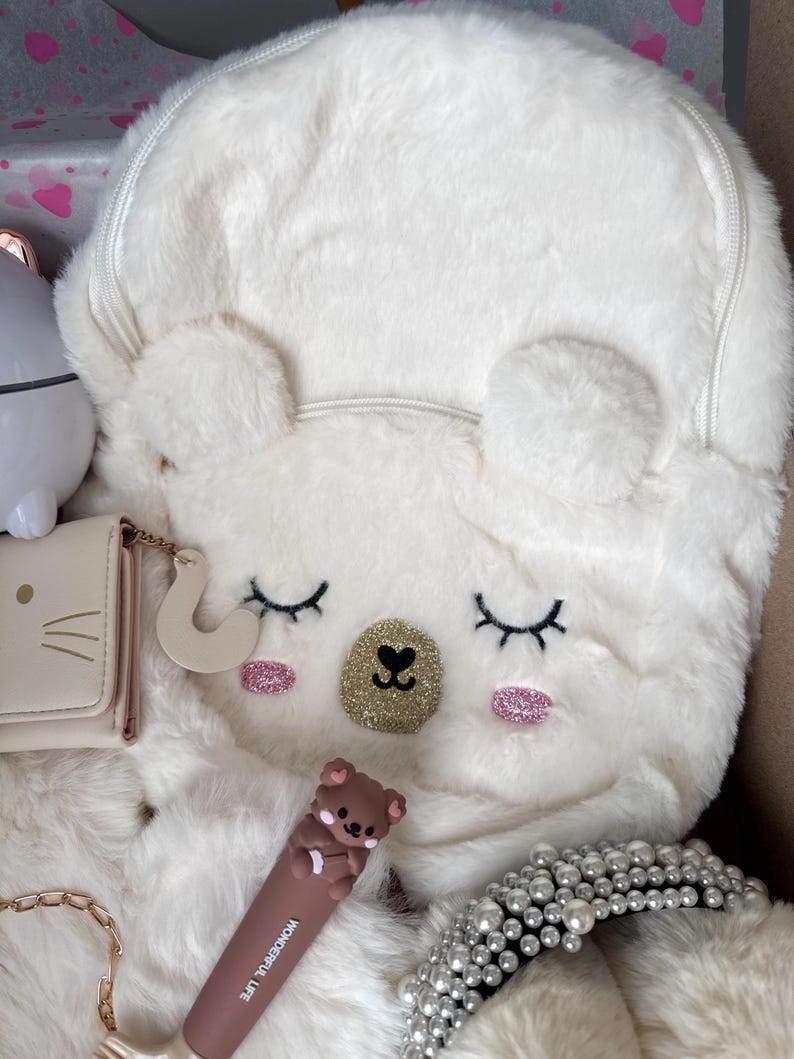 Puede incluir: Una mochila blanca y esponjosa con un dise&ntilde;o de cara de oso, con ojos cerrados, detalles de purpurina rosa y una nariz de purpurina dorada. Otros art&iacute;culos incluyen una cartera peque&ntilde;a, un labial marr&oacute;n con dise&ntilde;o de oso y una diadema de perlas.