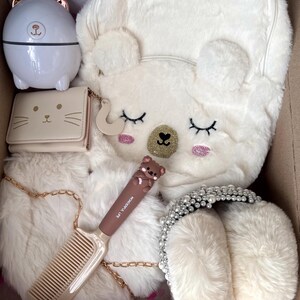 Puede incluir: Una colecci&oacute;n de accesorios blancos y beige, que incluye una mochila con orejas de oso, una cartera con cara de gato, un peine, una diadema y un peque&ntilde;o recipiente con forma de oso. Los art&iacute;culos est&aacute;n dispuestos en una caja de cart&oacute;n.