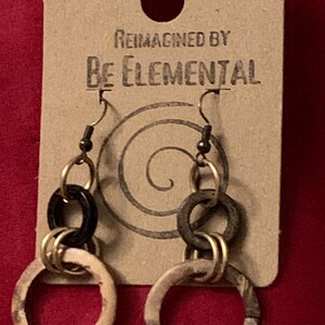Può includere: Orecchini dal design unico, con più anelli collegati in nero, oro e beige fantasia. Gli orecchini sono appesi a un gancio in metallo scuro. L'etichetta recita "Reimagined by Be Elemental".