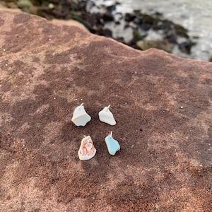 Può includere: Quattro piccoli ciondoli di forma irregolare in vetro marino, ciascuno con un'ansa color argento. I ciondoli sono in varie tonalità di bianco, blu e un motivo arancione e bianco. I ciondoli sono appoggiati su una superficie rocciosa marrone rossiccio.