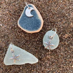 Puede incluir: Tres colgantes de cristal de mar con abalorios de plata. Un colgante es de color azul verdoso con un abalorio de media luna plateada. Los otros dos colgantes son de color verde claro con abalorios de estrella de mar plateados. Los colgantes están sobre una superficie arenosa.