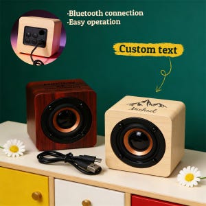 May include: Two wooden cube-shaped speakers, one in a dark wood grain finish and the other in a light wood finish. Each speaker has a black speaker cone with a copper-colored ring. The light wood speaker has the text "Michael" and a mountain graphic. The dark wood speaker has the text "ANDREW GROOMSMAN".