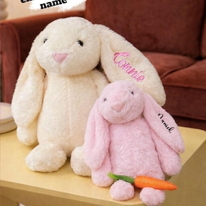 May include: Two plush bunny rabbits, one cream-colored and one pink, sit on a light yellow table. The cream bunny has "Connie" embroidered on its ear. The pink bunny has "Annid" embroidered on its ear and holds a plush carrot. The text "Custom embroidered name" is above the bunnies.