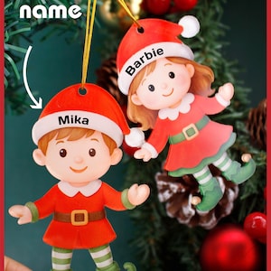 Può includere: Due decorazioni natalizie a forma di elfi, una con l'etichetta "Mika" e l'altra "Barbie". Gli elfi indossano abiti rossi con finiture bianche, scarpe verdi e cinture marroni. Lo sfondo presenta un albero di Natale con ornamenti rossi.