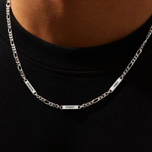 May include: A silver necklace featuring a figaro chain and three rectangular nameplates. The nameplates are engraved with the names "BRANDON", "ROBERT", and "JAMES". The necklace is shown against a black background.