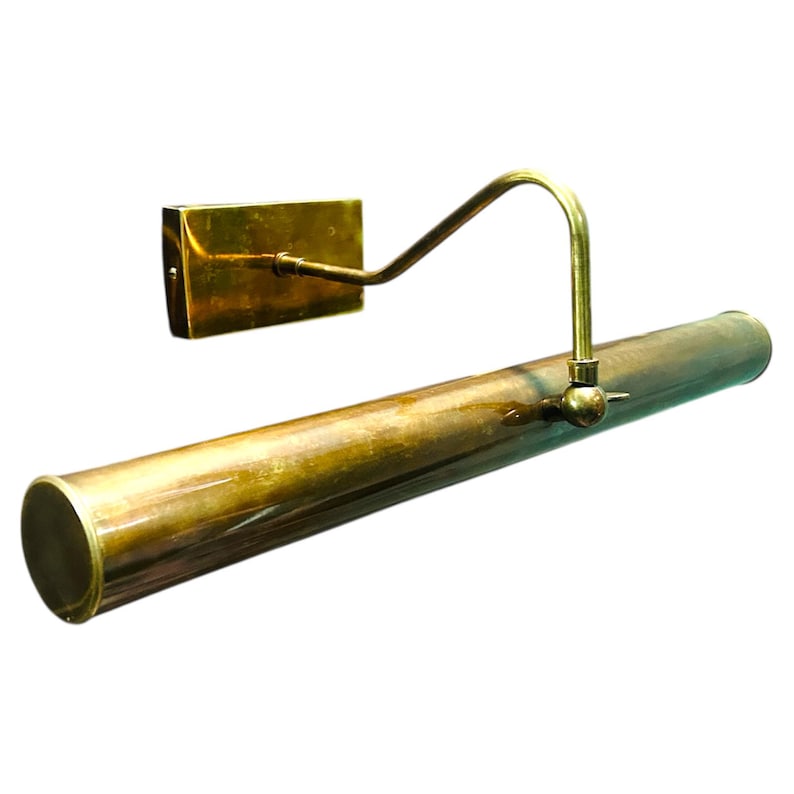 May include: A vintage-style brass picture light fixture. The fixture has a rectangular wall mount, a curved arm, and a long, cylindrical light housing. The brass has a slightly aged, tarnished appearance.