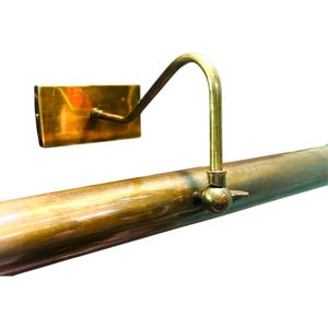 May include: A vintage-style brass picture light. The light features a rectangular wall mount, a curved arm, and a cylindrical light fixture. The brass has a warm, aged patina. The design is simple and elegant, suitable for illuminating artwork.