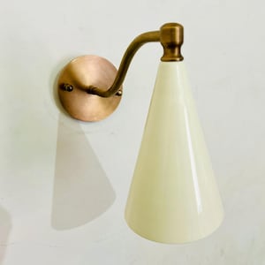 May include: A wall-mounted sconce with a cone-shaped, off-white shade. The fixture features a brass-colored curved arm and a circular copper-toned backplate. The sconce is designed for indoor lighting and adds a touch of vintage style.