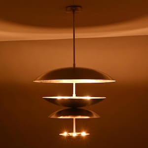Może przedstawiać: Nowoczesna lampa wisząca z trzema okrągłymi, metalowymi kloszami. Lampa zawieszona jest pod sufitem za pomocą cienkiego pręta. Każdy klosz posiada źródło światła, tworząc ciepłą, zachęcającą poświatę. Stylowy dodatek do każdego pomieszczenia.