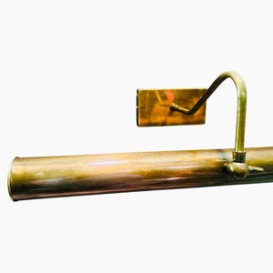 May include: A vintage brass picture light with a cylindrical body and a curved arm supporting a rectangular light fixture. The light has a warm, aged patina, suggesting it is an antique or a well-preserved piece.