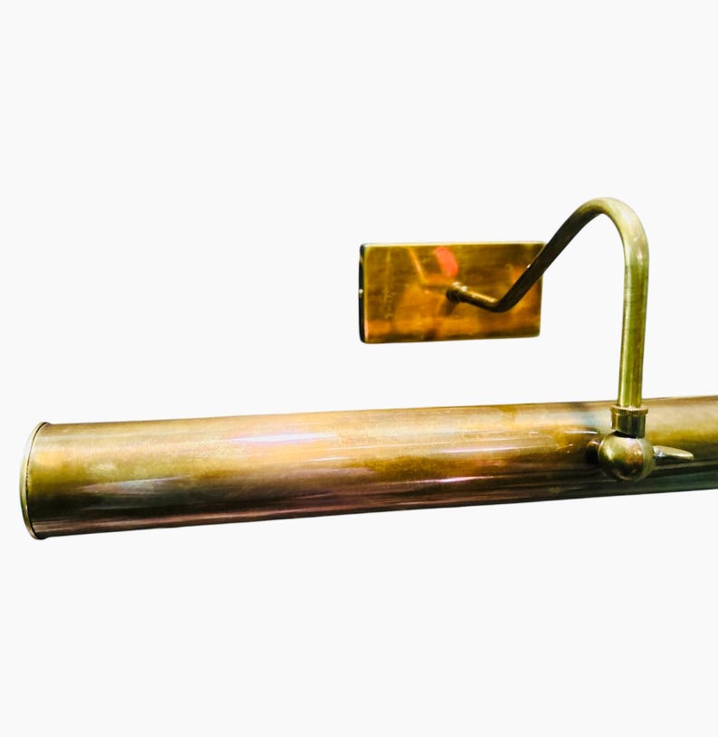 May include: A vintage brass picture light with a cylindrical body and a curved arm supporting a rectangular light fixture. The light has a warm, aged patina, suggesting it is an antique or a well-preserved piece.