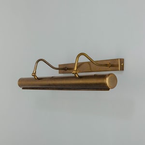 May include: A vintage-style picture light fixture with a brass finish. The fixture has a long, cylindrical body with two curved arms extending from it. The light is mounted on a rectangular backplate, designed to be mounted on a wall.