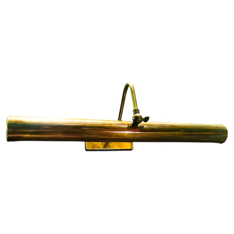 May include: A brass-toned picture light with a curved arm and adjustable head. The light is rectangular and mounted on a small, square base. The finish has a warm, antique appearance, suggesting a vintage or classic style.