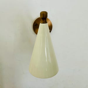 May include: Cream-colored cone-shaped wall sconce with a brass-colored mounting plate and a brass-colored top. The sconce is mounted on a white wall. The design is minimalist and modern.