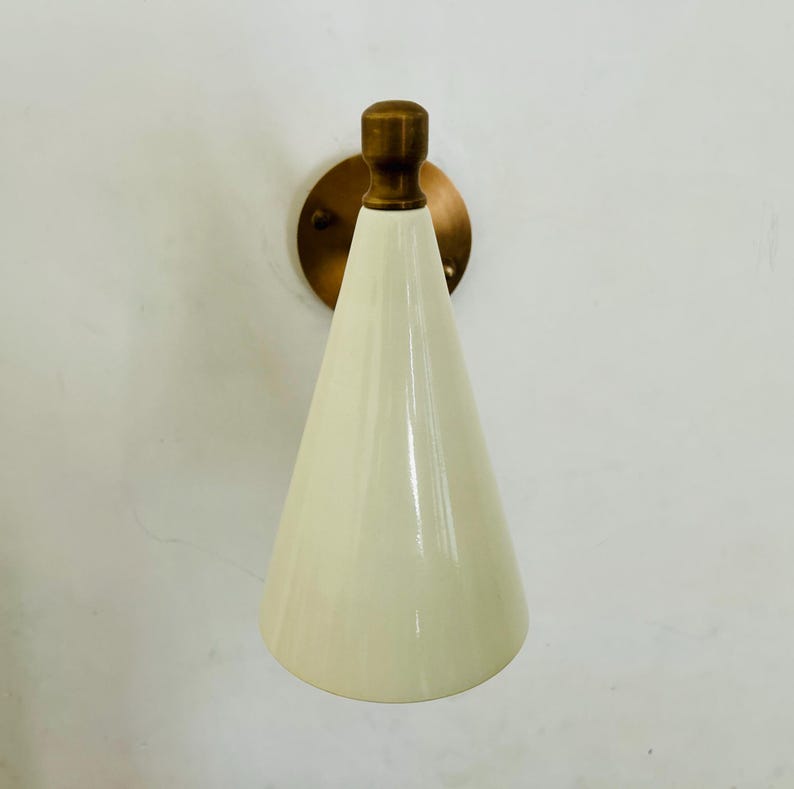 May include: Cream-colored cone-shaped wall sconce with a brass-colored mounting plate and a brass-colored top. The sconce is mounted on a white wall. The design is minimalist and modern.