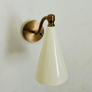 May include: A wall-mounted sconce with a brass finish and a conical, off-white shade. The sconce features a circular backplate and a curved arm connecting to the shade. The design is simple and elegant, suitable for various interior styles.