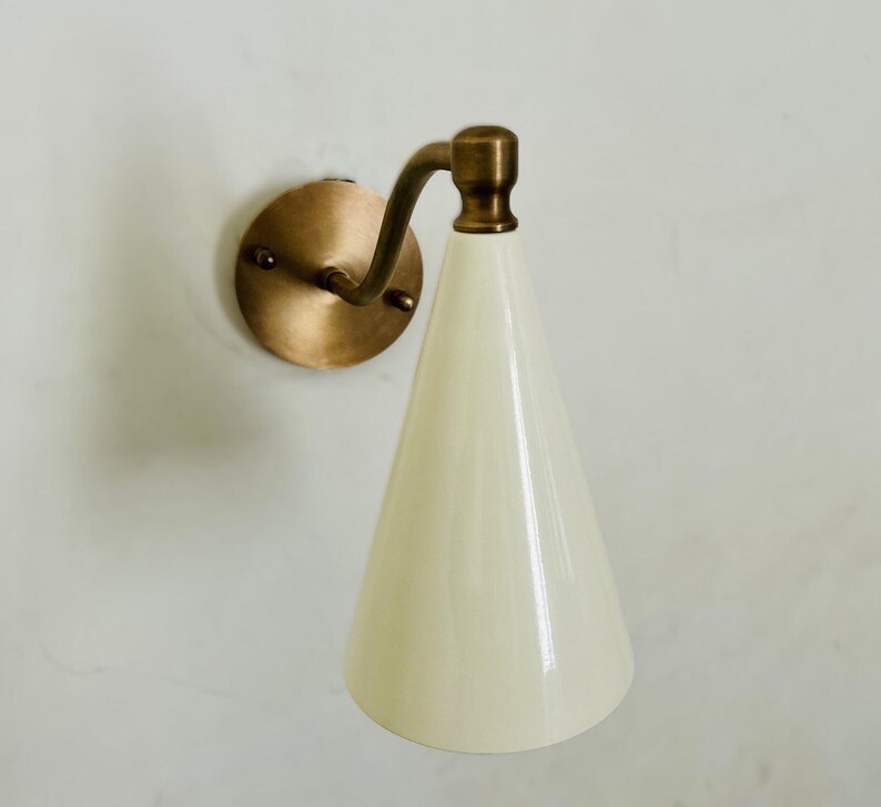 May include: A wall-mounted sconce with a brass finish and a conical, off-white shade. The sconce features a circular backplate and a curved arm connecting to the shade. The design is simple and elegant, suitable for various interior styles.