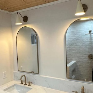 May include: Bathroom vanity with two arched mirrors, each framed in a gold finish. Above each mirror is a white cone-shaped light fixture. The countertop is white marble, and the sinks and faucets are gold.