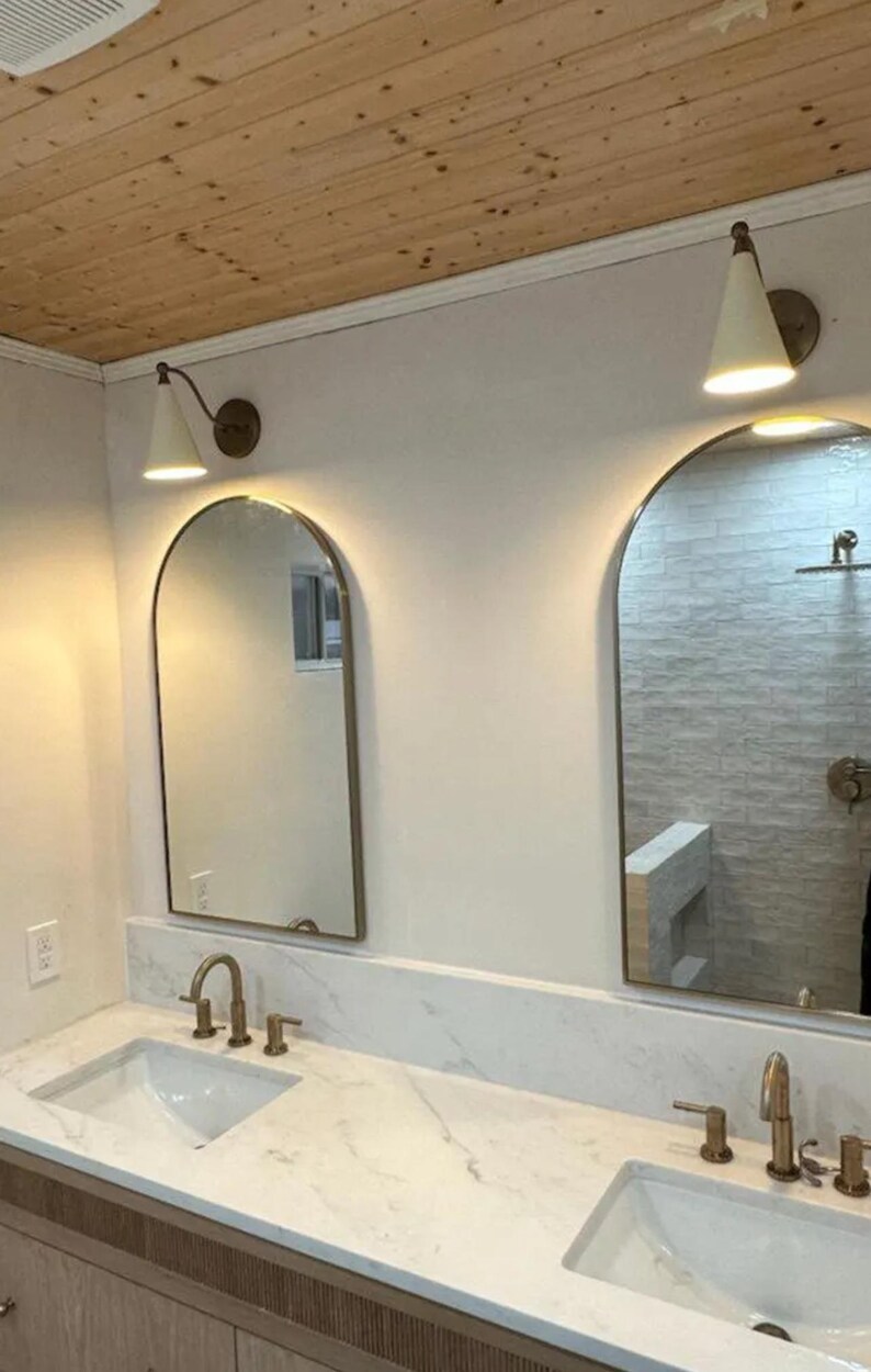 May include: Bathroom vanity with two arched mirrors, each framed in a gold finish. Above each mirror is a white cone-shaped light fixture. The countertop is white marble, and the sinks and faucets are gold.