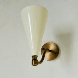 May include: A wall sconce with a cream-colored, cone-shaped shade and a brass-colored metal base. The sconce has a curved arm and a round backplate. The light fixture is mounted on a white wall.