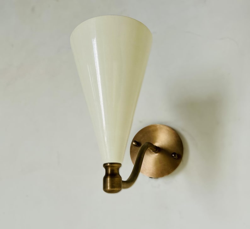 May include: A wall sconce with a cream-colored, cone-shaped shade and a brass-colored metal base. The sconce has a curved arm and a round backplate. The light fixture is mounted on a white wall.