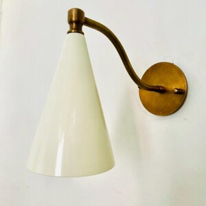 May include: A wall-mounted sconce with a conical, off-white shade and a curved brass arm. The sconce has a round brass base and a brass accent at the top of the shade. The design is mid-century modern.