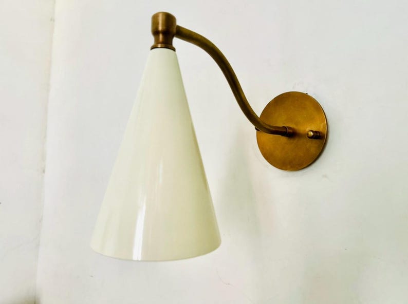May include: A wall-mounted sconce with a conical, off-white shade and a curved brass arm. The sconce has a round brass base and a brass accent at the top of the shade. The design is mid-century modern.