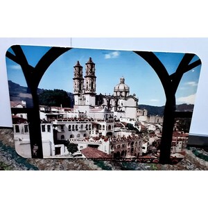 May include: A rectangular souvenir featuring a scenic view of a city with white buildings, red tile roofs, and a large church with two bell towers and a dome. The image is framed by a black arch against a blue sky.