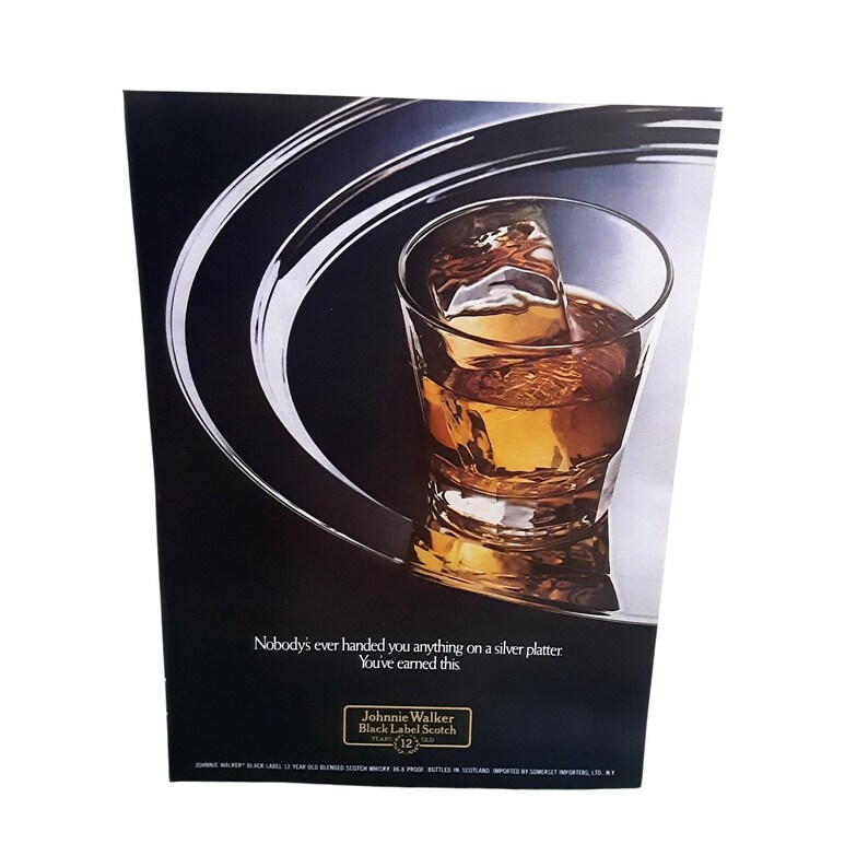 May include: An advertisement featuring a glass of amber-colored scotch with a large ice cube, set against a dark background with silver accents. The text reads, "Nobody's ever handed you anything on a silver platter. You've earned this." The Johnnie Walker Black Label logo is also visible.