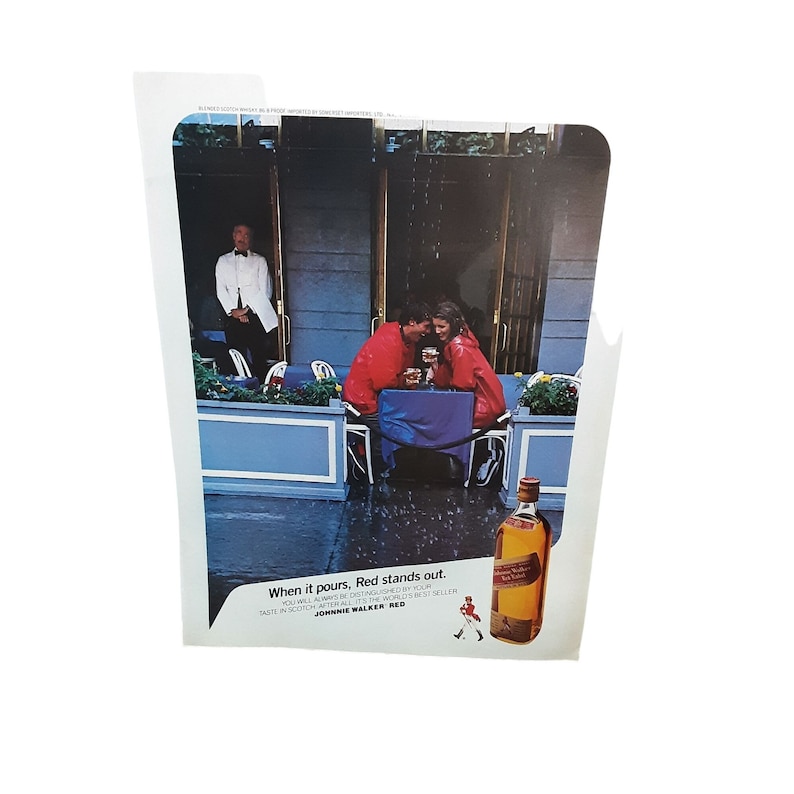 May include: A vintage advertisement for Johnnie Walker Red Label Scotch Whisky. The ad features a couple sitting at a table outside a cafe, enjoying a drink. The text reads "When it pours, Red stands out. You will always be distinguished by your taste in Scotch. After all, it's the world's best seller. Johnnie Walker Red."
