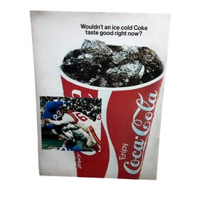 May include: Vintage Coca-Cola advertisement featuring a large red cup filled with ice and a dark beverage. The text reads, "Wouldn't an ice cold Coke taste good right now?" A small image of a football game is included.