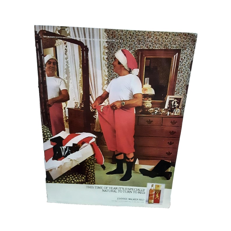 May include: A vintage advert showing a person in a Santa hat, white t-shirt, and red trousers, looking in a mirror. The text reads "THIS TIME OF YEAR IT'S ESPECIALLY NATURAL TO TURN TO RED" with "JOHNNIE WALKER RED" branding.