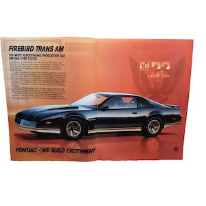 May include: A glossy black Pontiac Firebird Trans Am car ad. The ad features the text "FIREBIRD TRANS AM" and "THE MOST AERODYNAMIC PRODUCTION CAR GM HAS EVER TESTED". The car is parked on a red and orange background. The text "PONTIAC WE BUILD EXCITEMENT" is also visible.