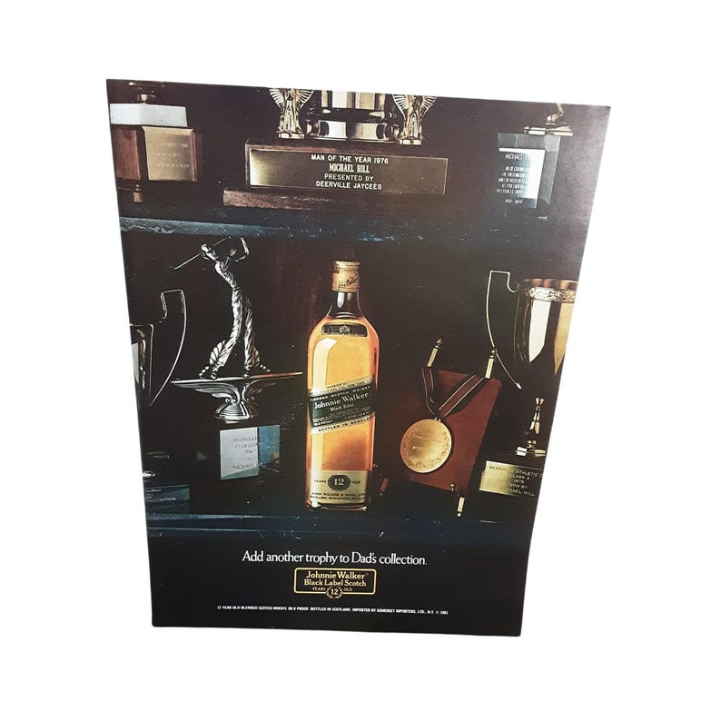 May include: An advertisement featuring a bottle of Johnnie Walker Black Label Scotch whisky, displayed among trophies and awards. The text reads "Add another trophy to Dad's collection."