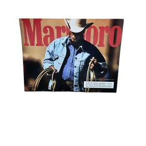 May include: A vintage Marlboro advertisement featuring a cowboy in a denim jacket and cowboy hat, holding a rope. The word "Marlboro" is in large red letters at the top. The ad includes a Surgeon General's warning at the bottom.