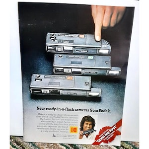 May include: Three black Kodak Ektralite cameras with built-in flash. The cameras are stacked on top of each other. The top camera has a 40 printed on it, the middle camera has a 20 printed on it, and the bottom camera has a 20 printed on it. The text "New, ready-in-a-flash cameras from Kodak." is printed below the cameras. A man is holding a camera and smiling. The text "Kodak gifts say "Open me first" to save Christmas in pictures" is printed below the man.