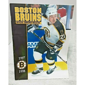 May include: A Boston Bruins official souvenir magazine from 1997-1998. The cover features a hockey player in a white, black, and gold uniform with the number 21. The magazine title is in yellow and black, with the Bruins logo.