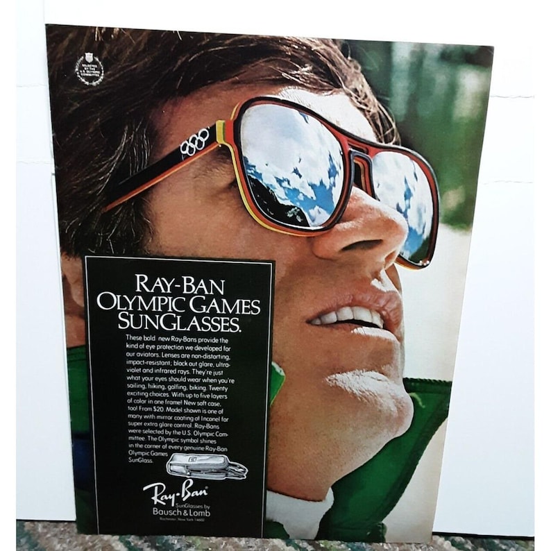 May include: A vintage advertisement for Ray-Ban Olympic Games sunglasses. The ad features a man wearing the sunglasses, which have a red and yellow frame and mirrored lenses. The text on the ad reads "Ray-Ban Olympic Games Sunglasses."