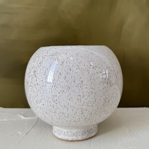 May include: A white ceramic vase with a speckled pattern. The vase has a rounded body and a short, pedestal base. The speckled design features small, brown dots across the white surface. The vase is a decorative item.