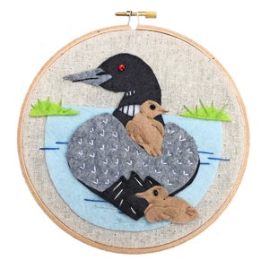 Puede incluir: Un aro de bordado de fieltro que representa un colimbo con dos polluelos. El colimbo es negro y gris con un ojo rojo. Los polluelos son marrones. El fondo es azul con hierba verde.
