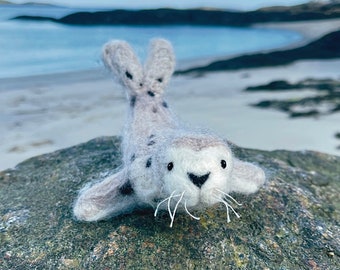 May include: A hand-crafted gray and white felt seal with black eyes and a white nose. The seal is lying on a gray rock with a blurred background of a beach and ocean.