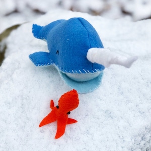 May include: A blue felt narwhal with a white horn and a red felt octopus on a white snowy background.