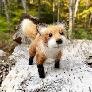 May include: A hand-felted fox figurine with a brown and white fur pattern, black eyes, and black feet. The fox is standing on a light brown, textured surface.