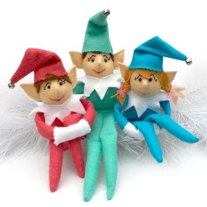 May include: Three felt elf dolls, one in red, one in teal, and one in blue, with white accents and embroidered faces. The elves are sitting on a white fluffy surface.