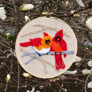 May include: A felt embroidery hoop with two red cardinals perched on a branch. The cardinals are facing each other and have white accents on their chests. The background is a beige fabric with a gray branch.