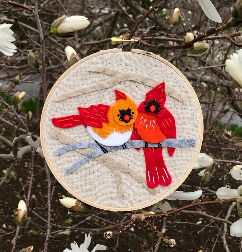 May include: A felt embroidery hoop with two red cardinals perched on a branch. The cardinals are facing each other and have white accents on their chests. The background is a beige fabric with a gray branch.