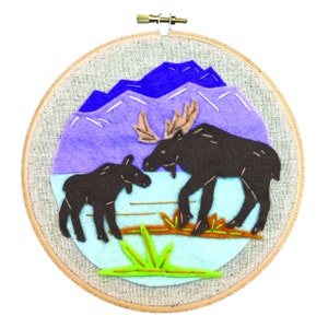 May include: A felt embroidery hoop with a blue mountain scene, a brown moose and a brown calf moose standing in a blue lake with green grass.