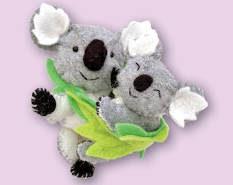 Può includere: Due orsi koala di feltro grigio che si abbracciano, uno più grande dell'altro, con foglie di feltro verde intorno a loro.