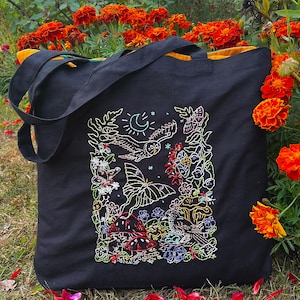 May include: Black tote bag with a colorful embroidered design featuring a moon, stars, birds, butterflies, mushrooms, and flowers. The bag has a yellow lining.